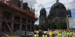 Bild von der Baustelle Berliner Schloss. Exkursionsteilnehmer laufen auf dem Gelände rum. Links Gebäude im Bauzustand. Geradeaus im Hintergrund ist die Kuppel des Schlosses zu sehen.