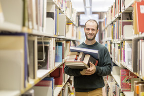 Ein Student steht in der Bibliothek im Gang und hat Bücher auf dem Arm.