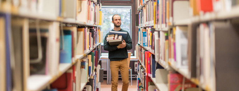 Student in der Bibliothek Studierender läuft an den Bücherregalen in der Bibliothek entlang.